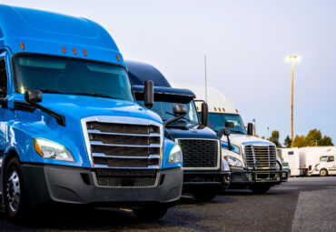 Different makes and models industrial grade professional big rigs semi trucks standing in row on the truck stop parking lot at evening time waiting to continued delivery schedule