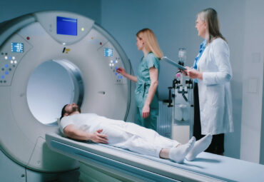 Young man lies down in MRI capsule. Chief head doctor in dressing gown hold folder and explains to patient what to do. Nurse monitors process of examiming. Man undergoes magnetic resonance imaging.