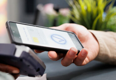 Female hand paying in clothing store with smartphone, tapping phone on contactless card reader while standing at cash register, close up. Customer using mobile phone as payment method in shopping mall